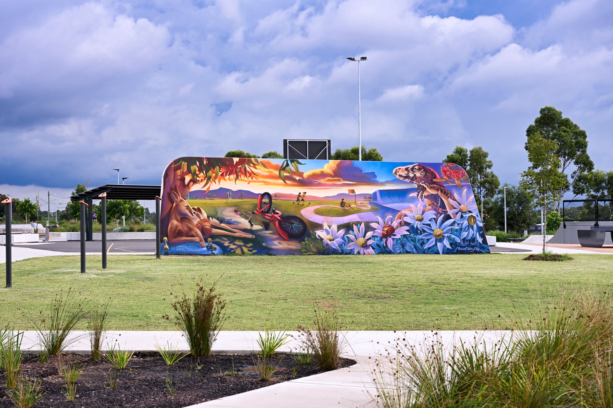A large mural at Gipps Street Recreation Precinct depicts a vibrant landscape with native plants, flowers and wildlife, including a lizard and kangaroo, alongside a bicycle and young people walking and riding. The mural sits within the recreation precinct