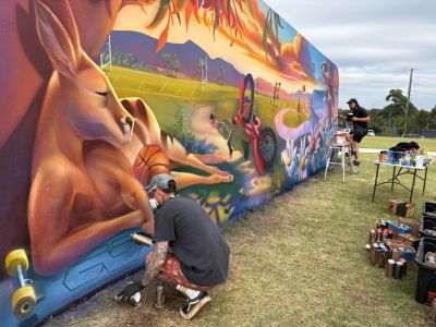 A large mural at Gipps Street Recreation Precinct depicts a vibrant landscape with native plants, flowers and wildlife, including a lizard and kangaroo, alongside a bicycle and young people walking and riding