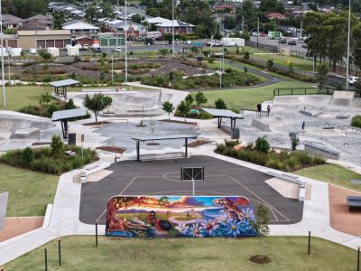 A large mural at Gipps Street Recreation Precinct depicts a vibrant landscape with native plants, flowers and wildlife, including a lizard and kangaroo, alongside a bicycle and young people walking and riding. The mural sits within the recreation precinct near the skate park and basketball facilities.